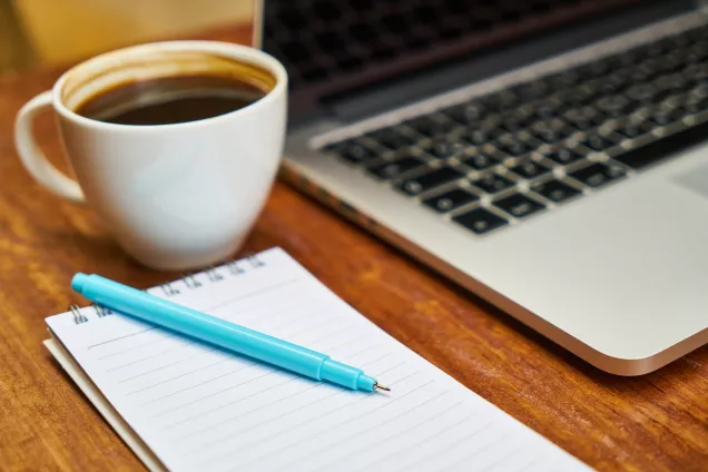 Bilden visar en kopp kaffe, en dator samt penna och block.
